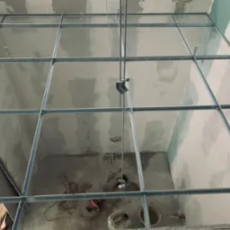 Metalowa konstrukcja sufitowa z profili, widok z dołu, częściowo obłożona płytami gipsowo-kartonowymi, widoczne narzędzia i pozostałości materiałów budowlanych na podłodze.