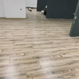 Nowo ułożone panele podłogowe imitujące drewno w jasnym odcieniu w biurze, widoczne przygotowania do dalszych prac wykończeniowych, ściany w kolorze białym i grafitowym.