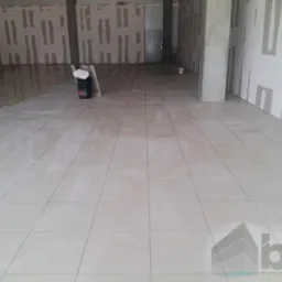 Świeżo położone, jasne płytki podłogowe w surowym wnętrzu biurowym z widocznymi śladami prac wykończeniowych na ścianach i wiaderkiem po kleju.