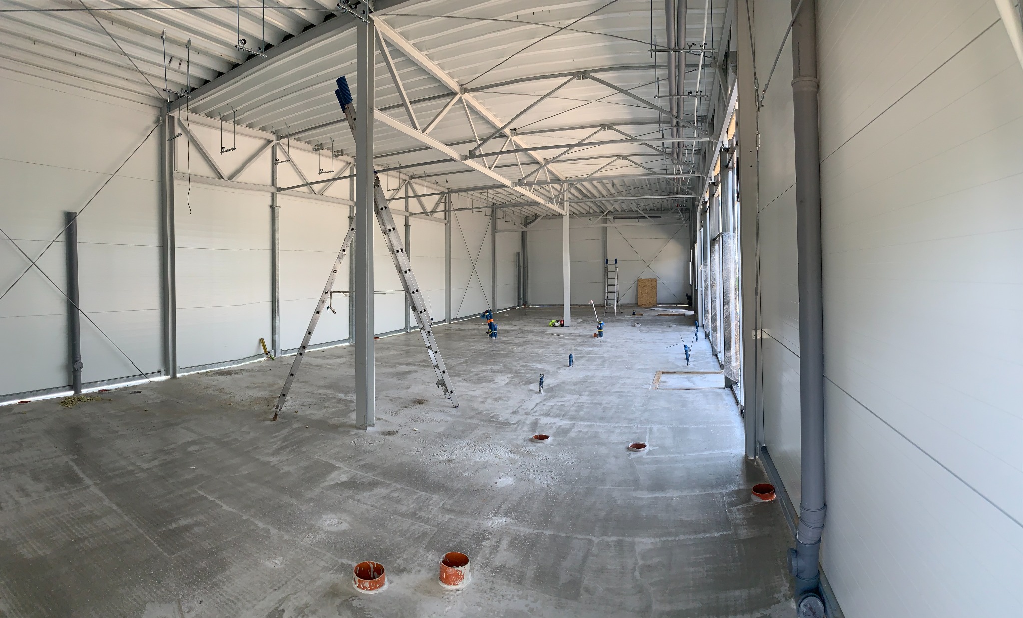 Wnętrze hali produkcyjnej w trakcie budowy, widoczne stalowe elementy konstrukcyjne, ściany z paneli, betonowa posadzka z instalacjami, drabina oparta o słup.