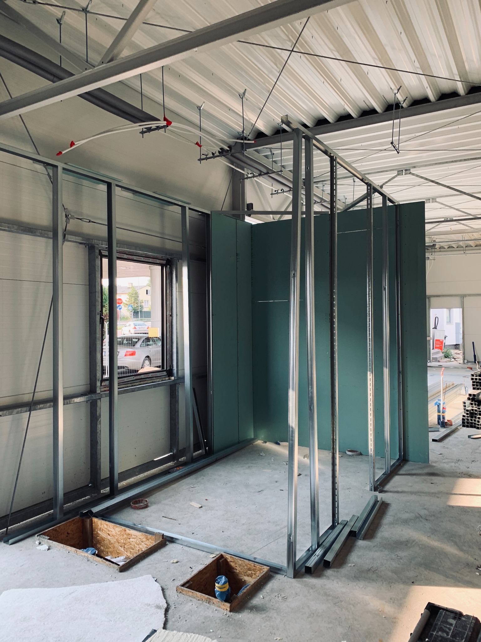 Montaż ścian działowych z płyt kartonowo-gipsowych w surowym wnętrzu hali, widoczne metalowe profile, częściowo zamontowane zielone płyty, otwory w podłodze i instalacja przeciwpożarowa pod sufitem.