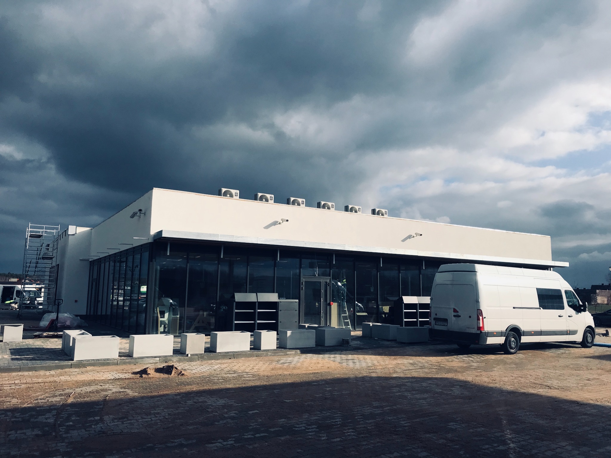 Nowo wybudowany, parterowy budynek biurowy z przeszkloną fasadą i rzędem klimatyzatorów na dachu, widoczny podczas prac wykończeniowych na zewnątrz, zaparkowany biały samochód dostawczy, pochmurne...