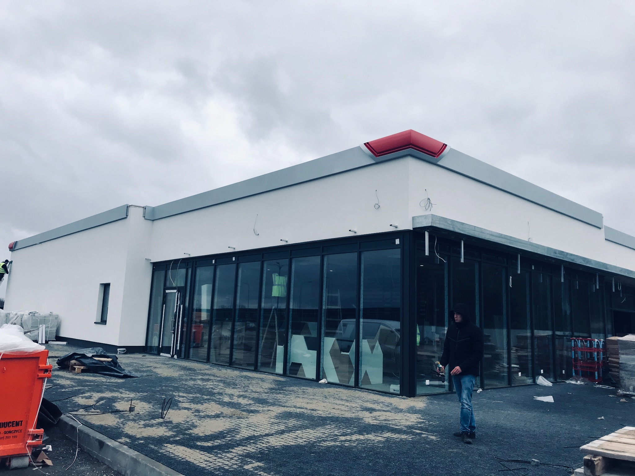 Nowo wybudowany, parterowy budynek komercyjny z dużymi oknami, widoczny mężczyzna w kurtce i jeansach, teren wokół w trakcie prac wykończeniowych, pochmurne niebo.