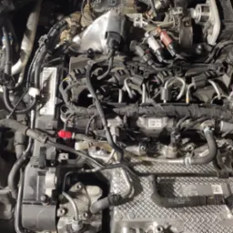 Widok z góry na skomplikowaną konstrukcję silnika samochodowego z widocznymi przewodami, wtryskiwaczami, pokrywą zaworów i innymi elementami osprzętu, oświetlonego sztucznym światłem.