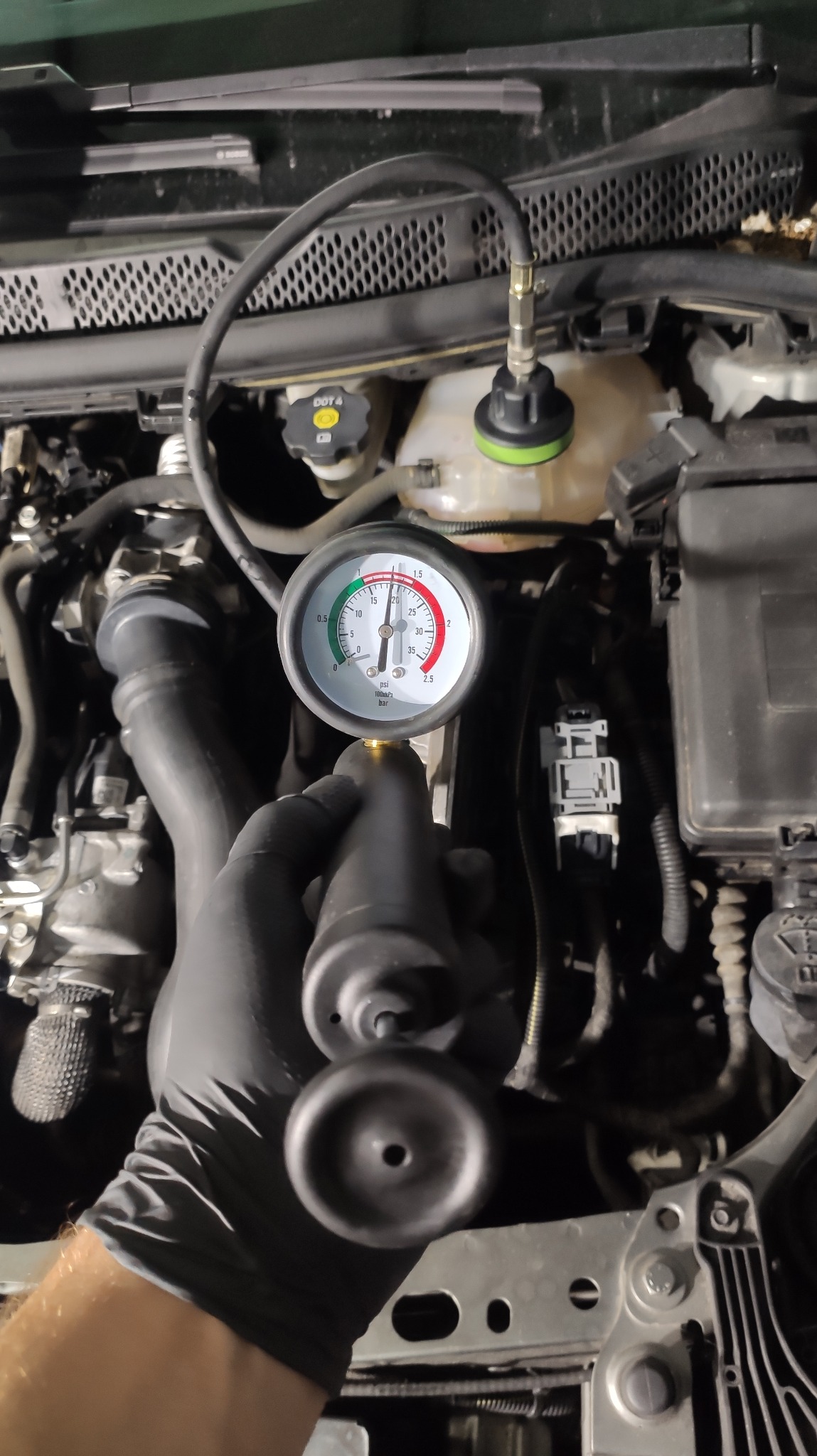 Sprawdzanie szczelności układu chłodzenia silnika samochodowego za pomocą testera ciśnieniowego; widoczny manometr wskazujący ciśnienie w układzie, podłączony do zbiornika wyrównawczego płynu...