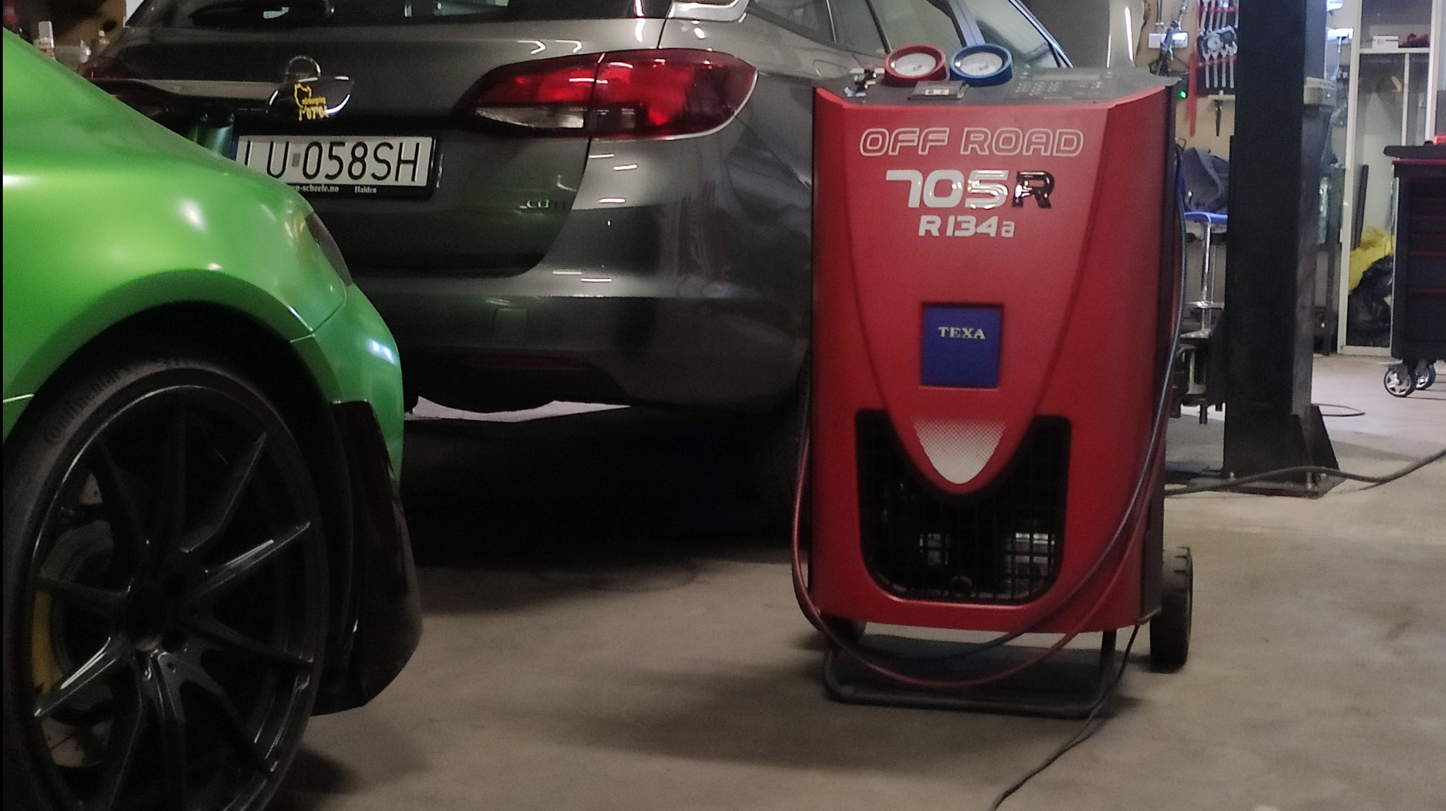 Czerwone urządzenie do obsługi klimatyzacji samochodowej TEXA 705R R134a stoi obok szarego samochodu Opel Astra i fragmentu zielonego auta w warsztacie.