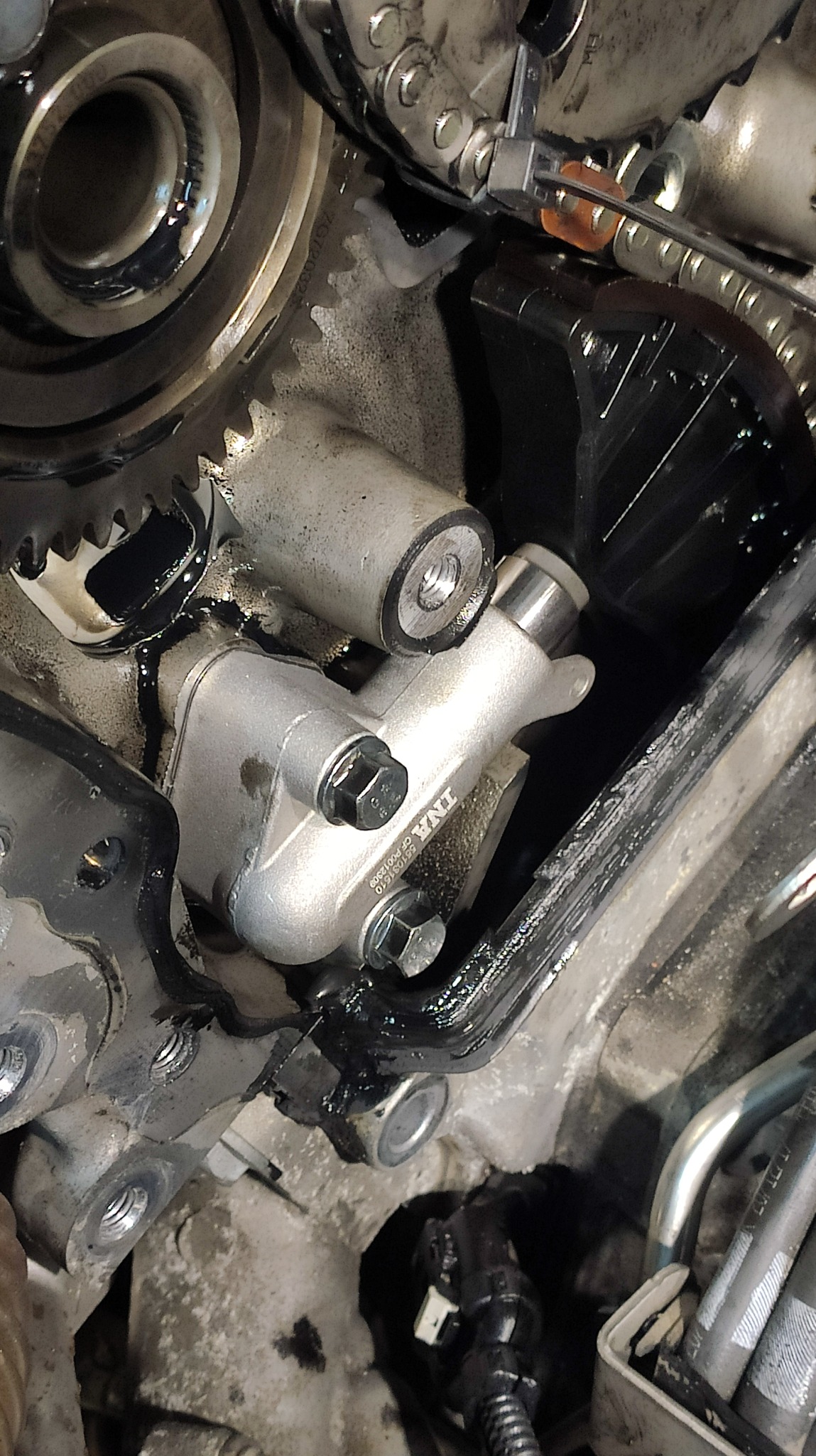 Szczegółowe zbliżenie na mechanizm rozrządu silnika samochodowego, z widocznym napinaczem łańcucha, kołami zębatymi i łańcuchem, wszystko pokryte świeżym olejem.