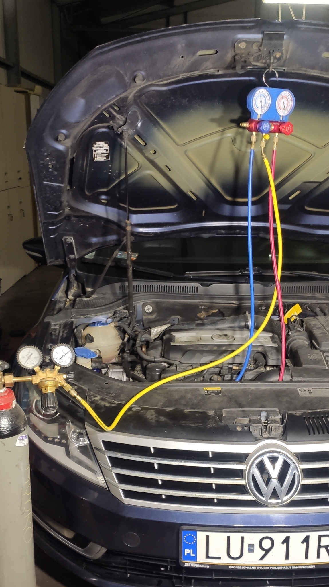 Niebieski Volkswagen Passat z otwartą maską, podłączony do manometrów klimatyzacji z kolorowymi przewodami, butla z czynnikiem chłodniczym po lewej stronie.