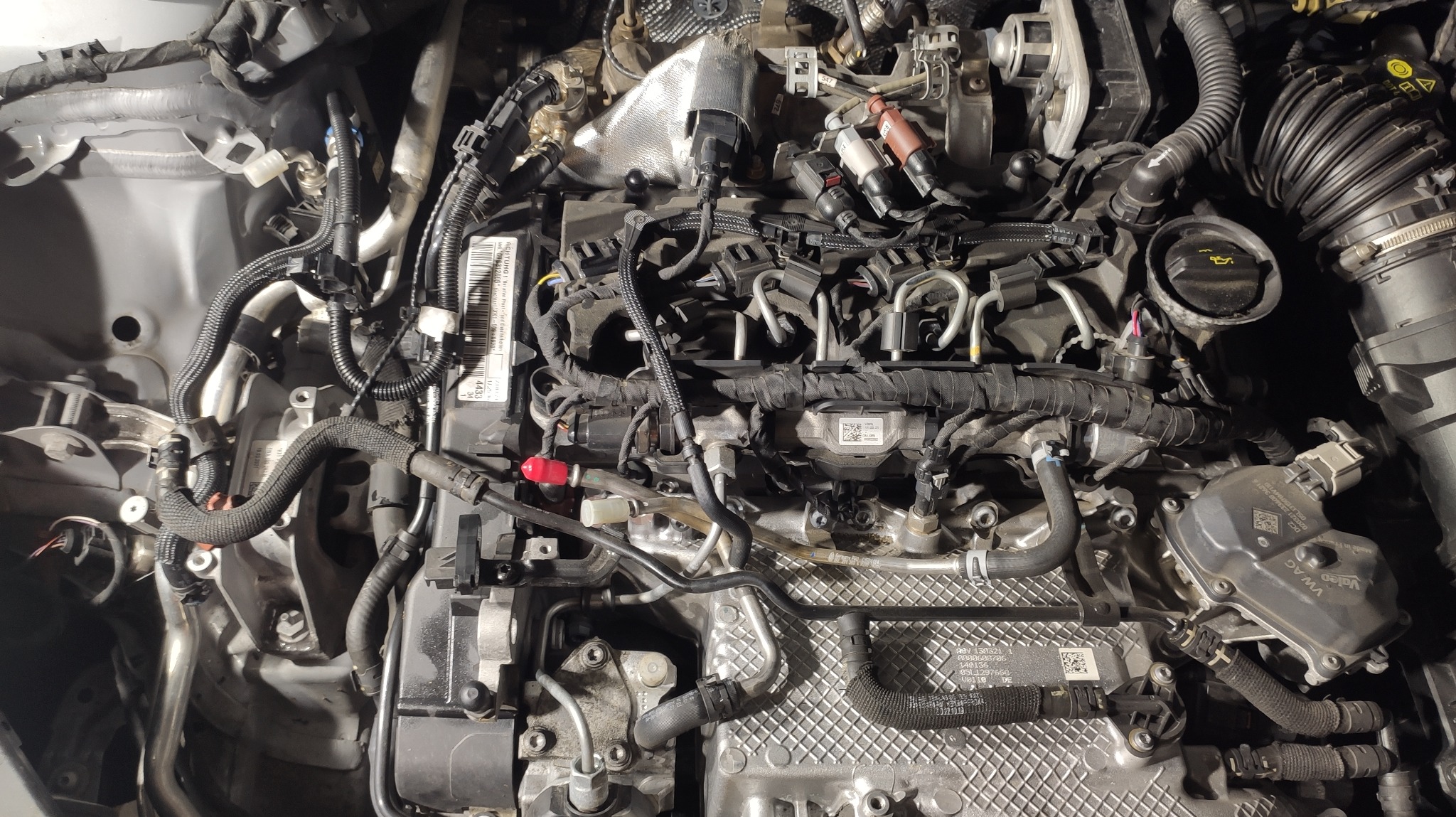 Widok z góry na skomplikowaną konstrukcję silnika samochodowego z widocznymi przewodami, wtryskiwaczami, pokrywą zaworów i innymi elementami osprzętu, oświetlonego sztucznym światłem.