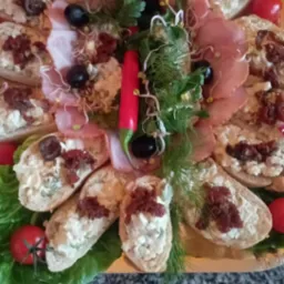 Dekoracyjna taca z kanapkami z pastą, szynką, oliwkami, suszonymi pomidorami, czerwoną papryczką chili i zielenią, na tle marmurowego blatu.