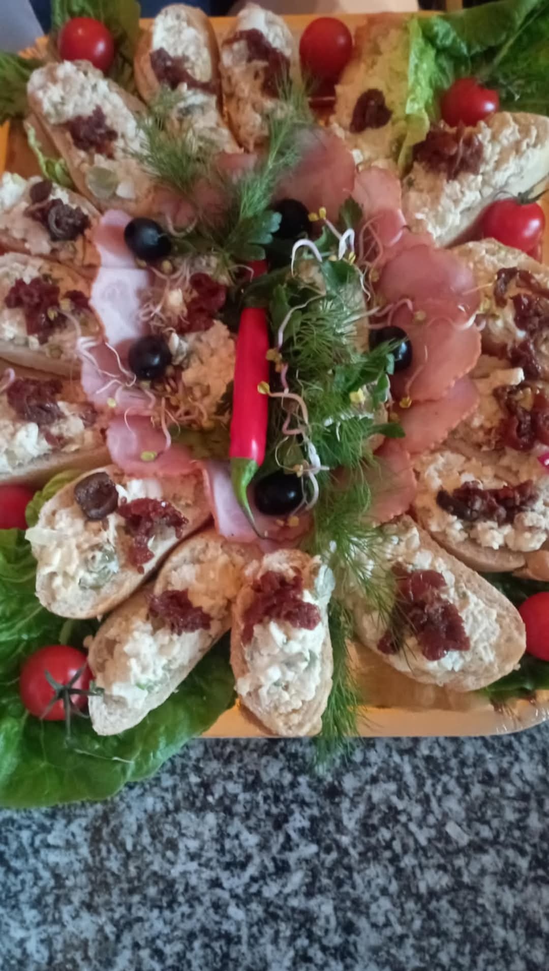 Dekoracyjna taca z kanapkami z pastą, szynką, oliwkami, suszonymi pomidorami, czerwoną papryczką chili i zielenią, na tle marmurowego blatu.