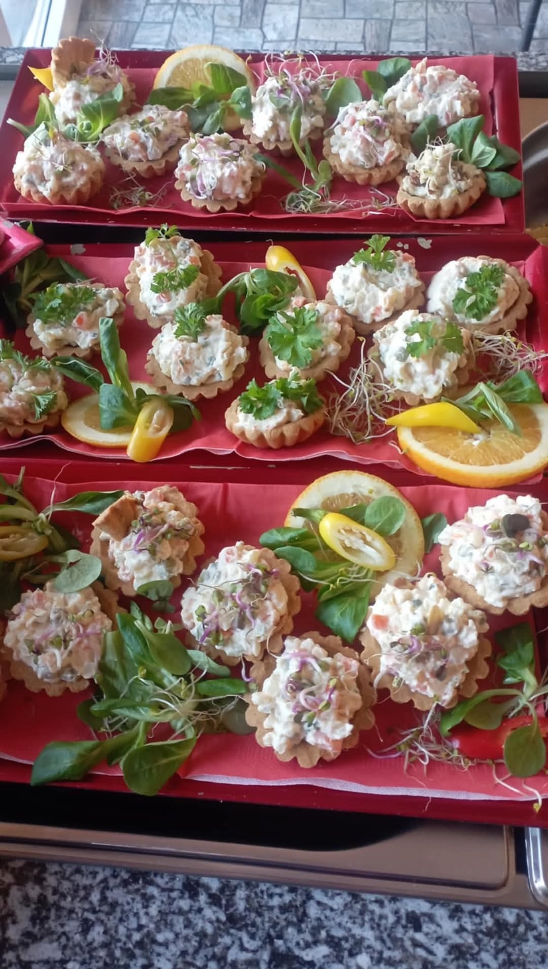 Tarty z kremowym farszem, dekorowane zieleniną i plasterkami cytrusów, ułożone na czerwonych tacach.