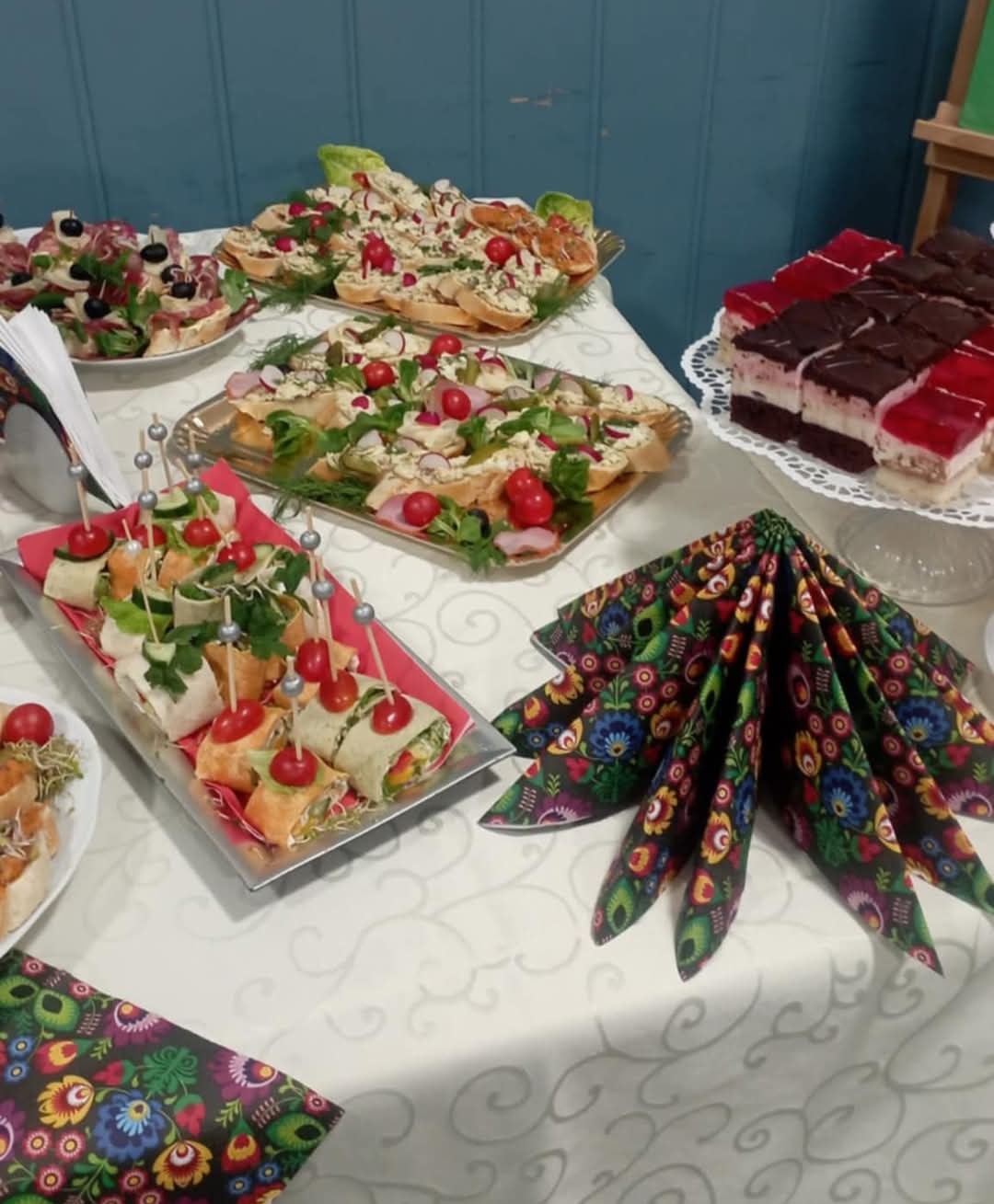 Stół z przekąskami: kanapeczki z warzywami i pastami, tortille z nadzieniem, ciasto w trzech kolorach na paterze, serwetki w ludowe wzory, biały obrus w delikatne wzory.