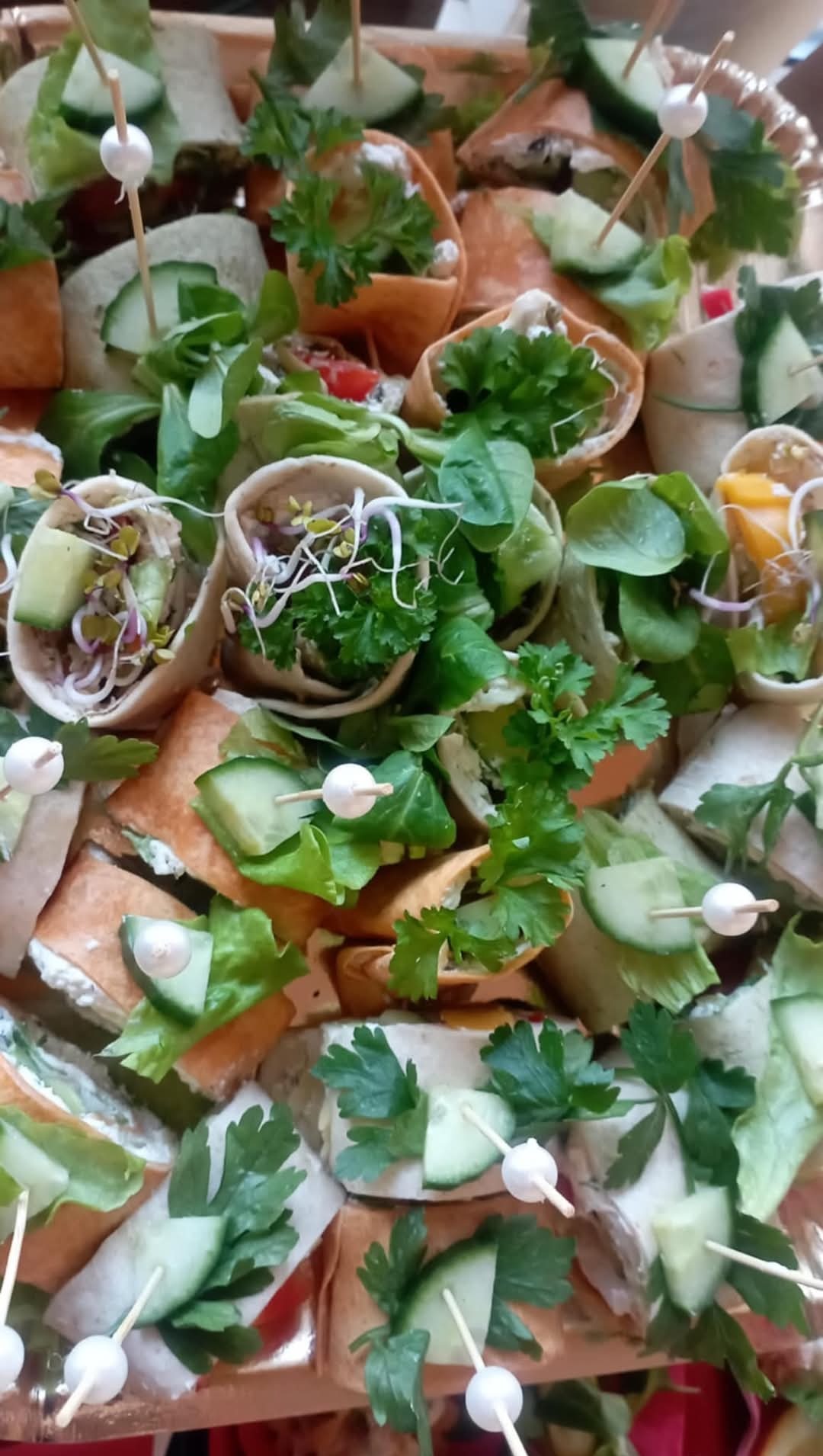 Różnorodne przekąski w tortillach i wrapach, udekorowane świeżymi ziołami, ogórkiem i kiełkami, spięte ozdobnymi wykałaczkami, tworzące kompozycję na przyjęcie.