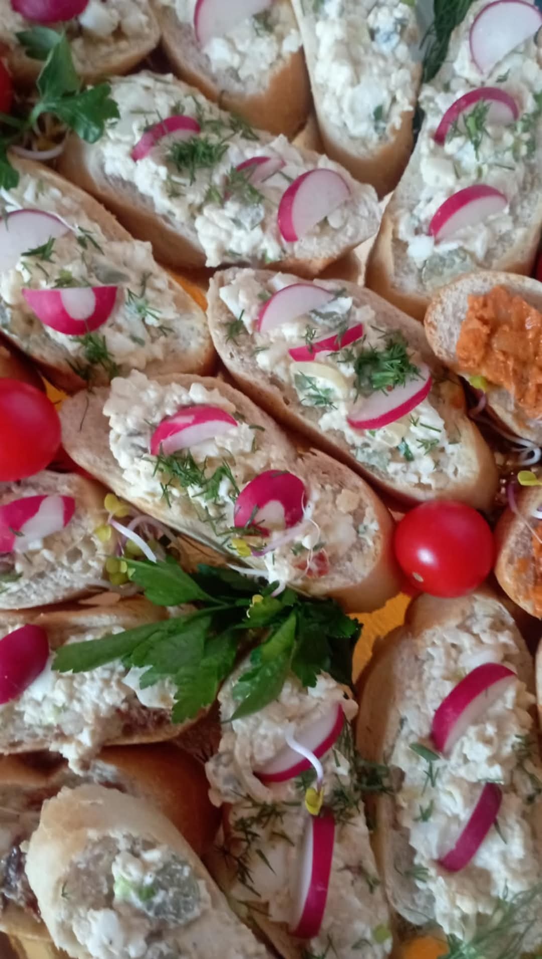 Koreczki z pastą jajeczną, rzodkiewką i koperkiem, ułożone na tacy z dodatkiem pomidorków koktajlowych i świeżych ziół.