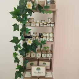 Półka z naturalnymi produktami konopnymi: szampony, mydła, kremy do twarzy, peelingi i susz, ozdobiona pnączem bluszczu i białymi kwiatami, eksponowana na kolumnie.