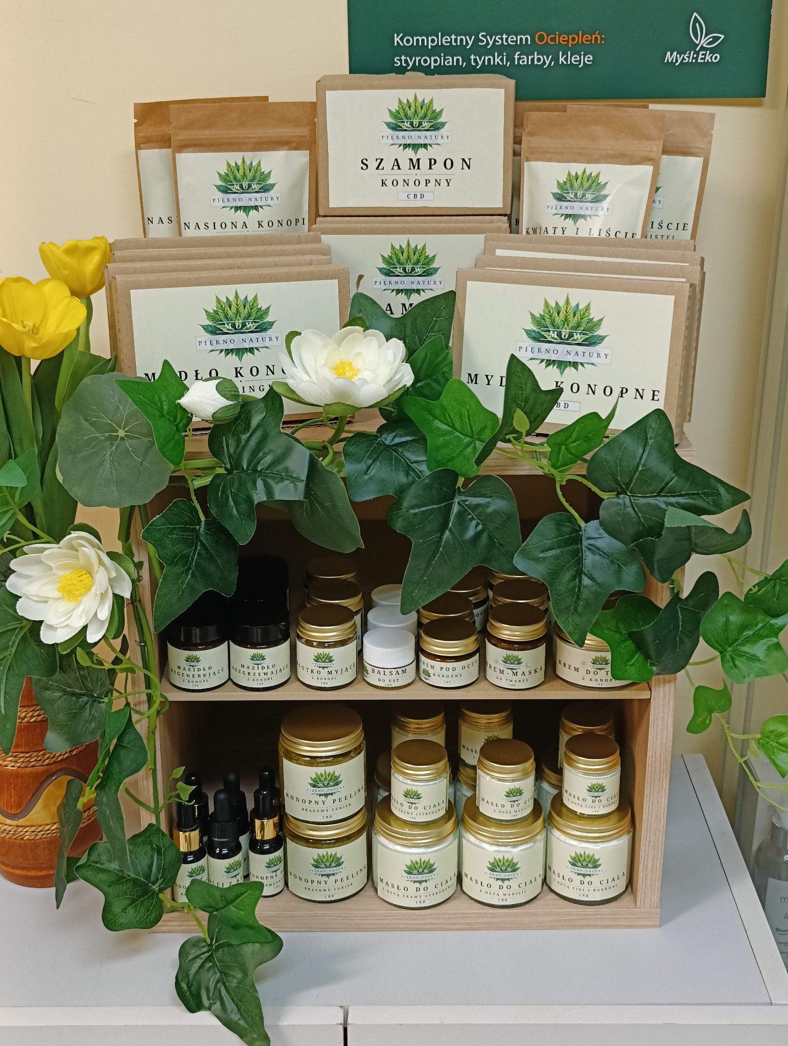 Półka z naturalnymi kosmetykami konopnymi marki Piękno Natury, w drewnianej skrzynce, z dodatkiem dekoracyjnych kwiatów i pnączy, na pierwszym planie olejki w ciemnych buteleczkach z pipetami,...