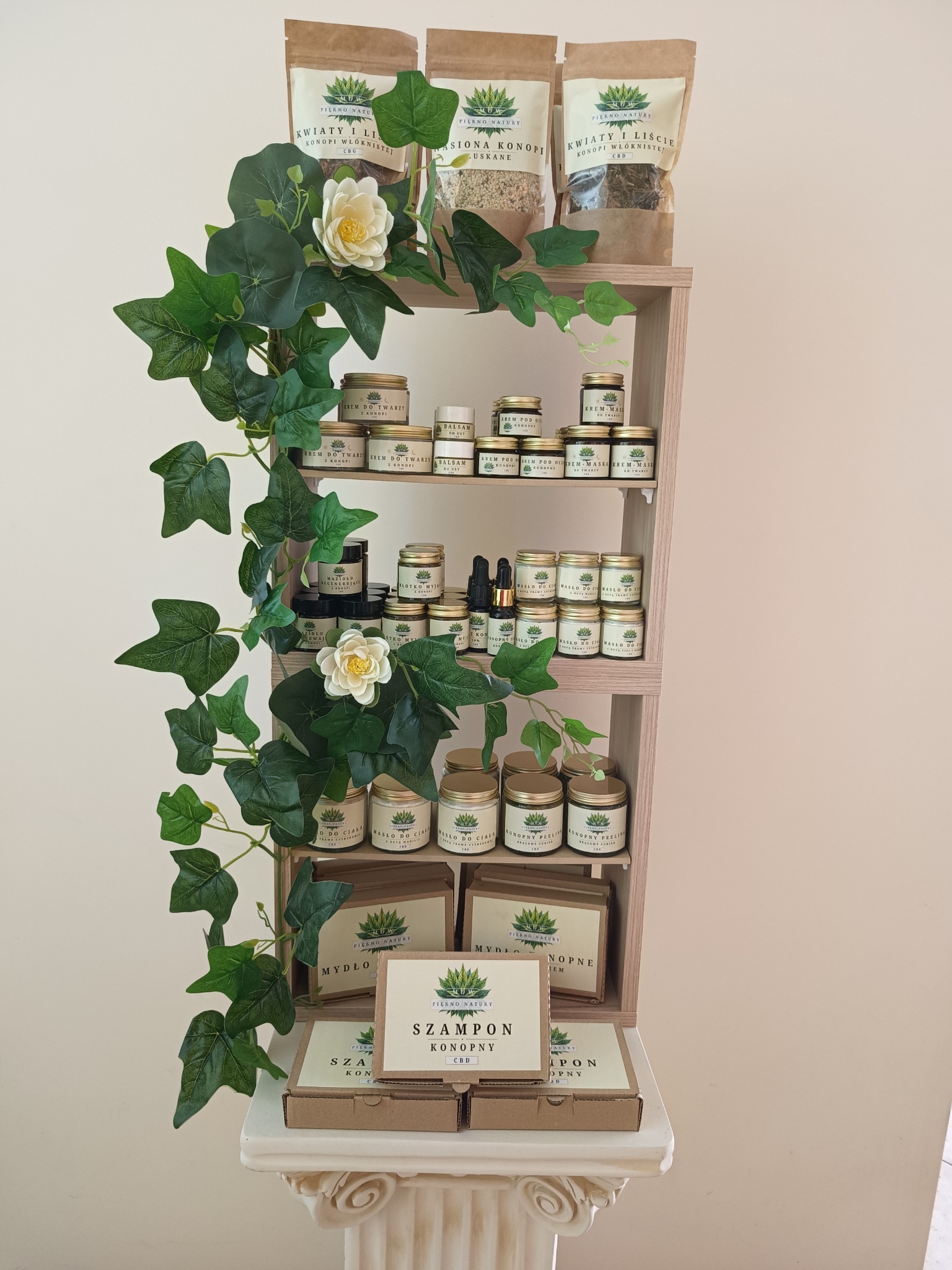 Półka z naturalnymi produktami konopnymi: szampony, mydła, kremy do twarzy, peelingi i susz, ozdobiona pnączem bluszczu i białymi kwiatami, eksponowana na kolumnie.