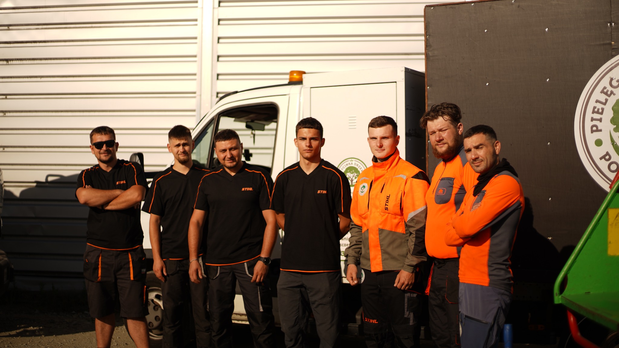 Grupa sześciu mężczyzn w strojach roboczych firmy Stihl pozuje na tle białego samochodu dostawczego i przyczepy z logo firmy oferującej pielęgnację drzew.