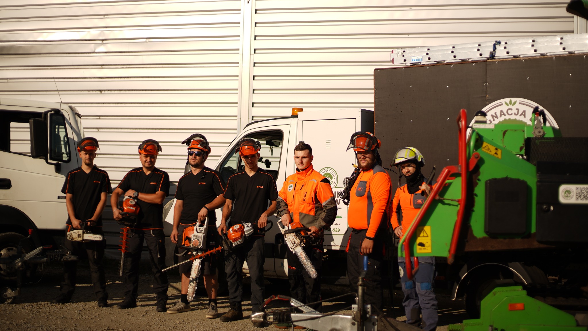 Grupa siedmiu pracowników w strojach roboczych Stihl, z kaskami i ochroną słuchu, pozuje z piłami łańcuchowymi i nożycami do żywopłotu na tle białego samochodu dostawczego i zielonej rębarki...