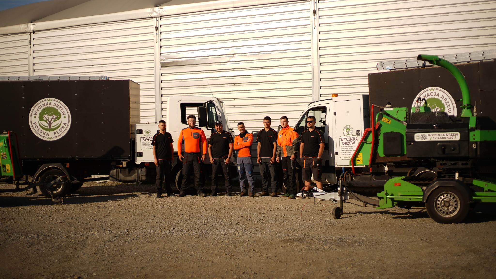 Zespół pracowników firmy zajmującej się wycinką drzew pozuje na tle pojazdów z logo firmy i rębakiem do gałęzi.