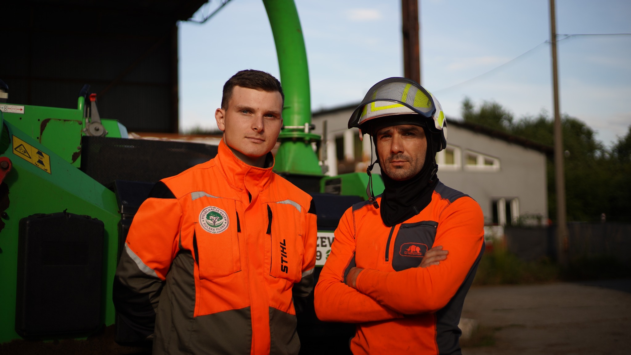 Dwóch mężczyzn w pomarańczowych uniformach ochronnych, jeden w kasku, pozuje przed zielonym rębakiem do gałęzi na tle budynku.