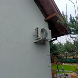 Biała jednostka zewnętrzna klimatyzatora z logo Rotenso zamontowana na szarej ścianie domu, widok z boku na tle iglastego drzewa i drabiny.