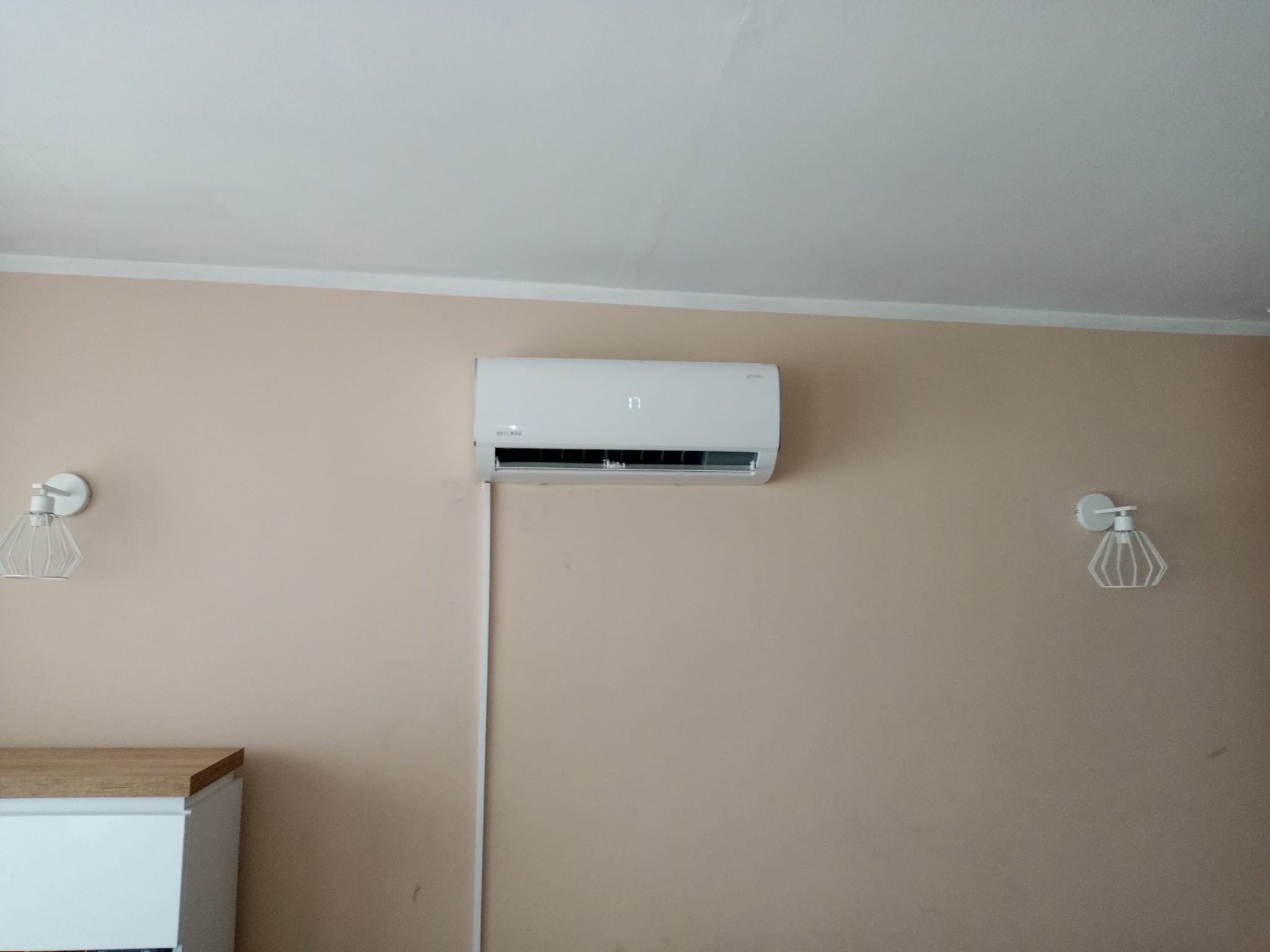 Biała jednostka klimatyzacji zamontowana na beżowej ścianie w pokoju, widoczne lampki nocne po bokach i fragment szafki.