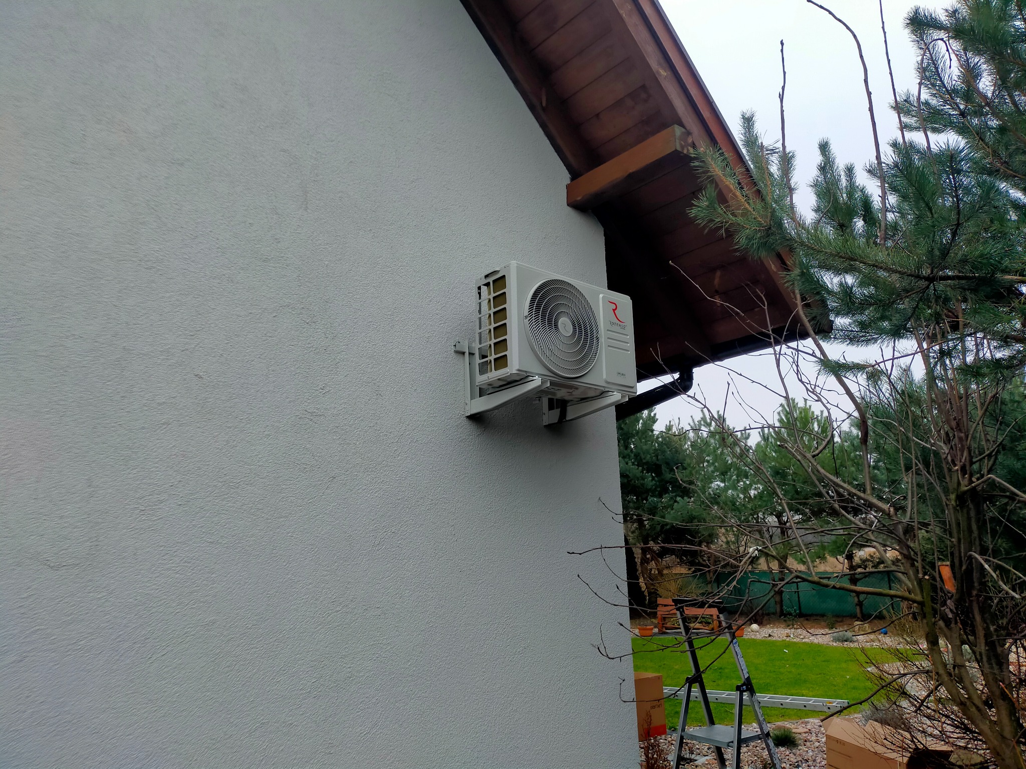 Biała jednostka zewnętrzna klimatyzatora z logo Rotenso zamontowana na szarej ścianie domu, widok z boku na tle iglastego drzewa i drabiny.