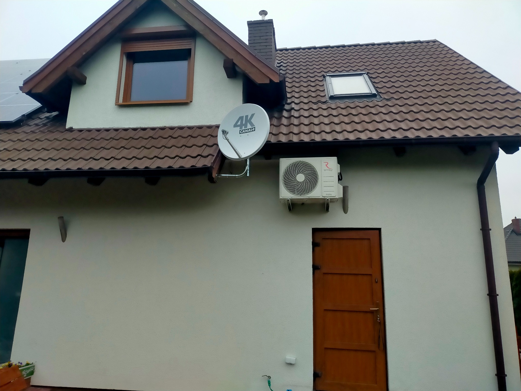 Biały budynek jednorodzinny z brązowym dachem, oknami i drzwiami, na ścianie zewnętrzna jednostka klimatyzacji oraz antena satelitarna.