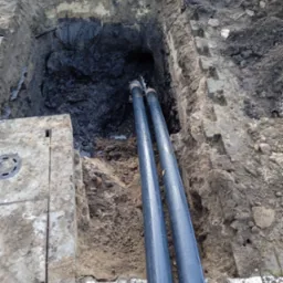 Dwie rury prowadzone w wykopie ziemnym, widoczny betonowy właz studzienki kanalizacyjnej po lewej stronie.