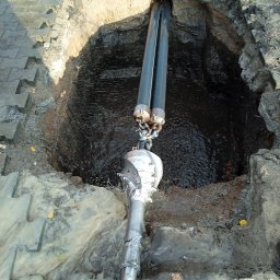 FIRMA HANDLOWO USŁUGOWA PRZEWIERTY STEROWANE-NOWI Robert Pujanek - Wiertnica kierunkowa w wykopie, widoczne dwie rury i głowica wiertnicza w błocie, otoczenie z betonowej kostki brukowej i ziemi.