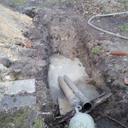 FIRMA HANDLOWO USŁUGOWA PRZEWIERTY STEROWANE-NOWI Robert Pujanek - Dwa rurociągi w wykopie wypełnionym wodą, z widocznym elementem wiertniczym i błotnistym otoczeniem, w tle tor kolejowy.