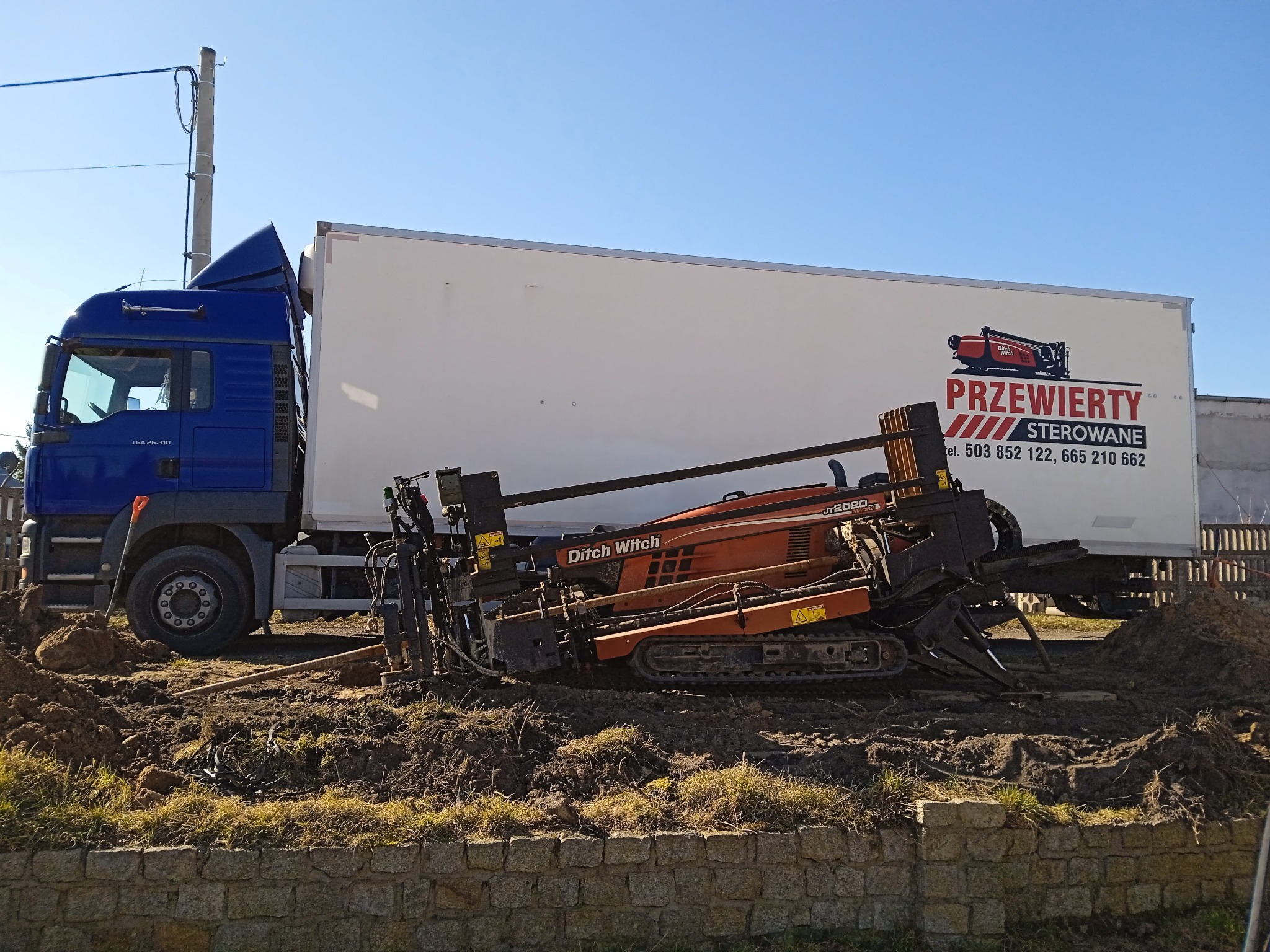 Pomarańczowa maszyna Ditch Witch JT2020 do robót ziemnych stoi na błotnistym terenie obok niebieskiej ciężarówki z białą naczepą z logo firmy. W tle błękitne niebo.