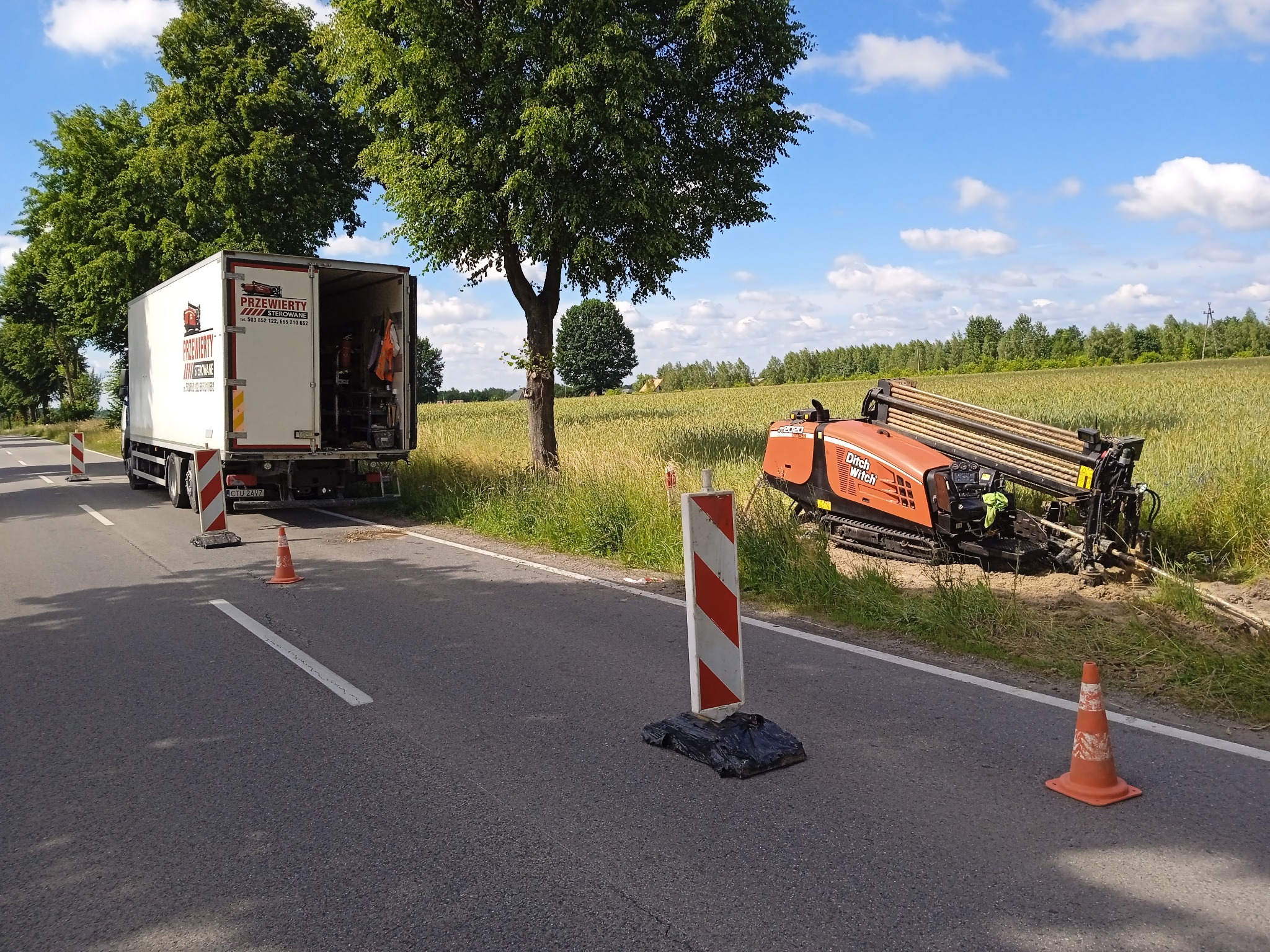 Pomarańczowa wiertnica Ditch Witch ustawiona na poboczu drogi, obok otwartej ciężarówki z logo firmy, w tle pole i drzewa.