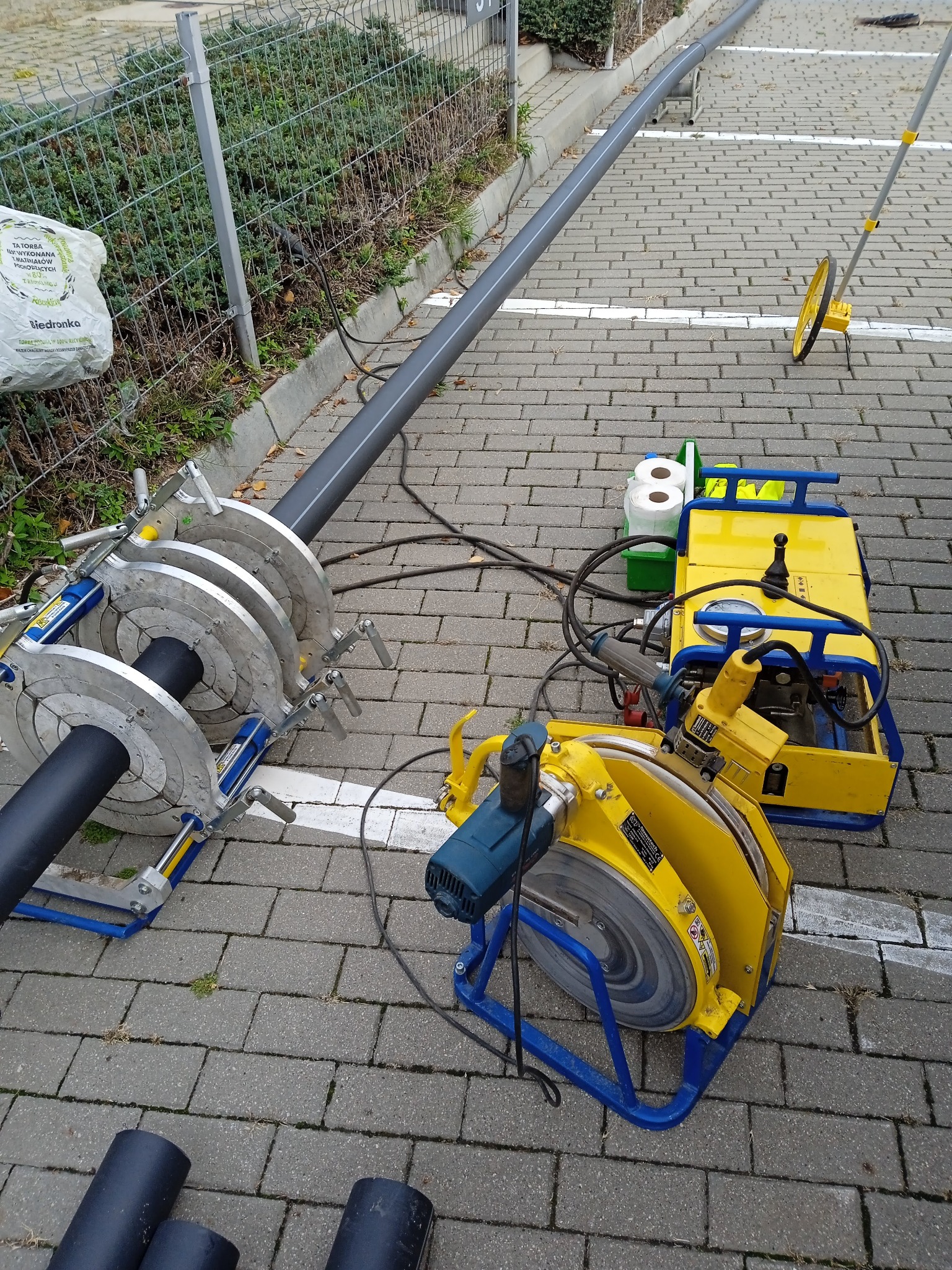 Maszyny do łączenia rur z tworzyw sztucznych ustawione na kostce brukowej, widoczna długa rura, obok torba z Biedronki.
