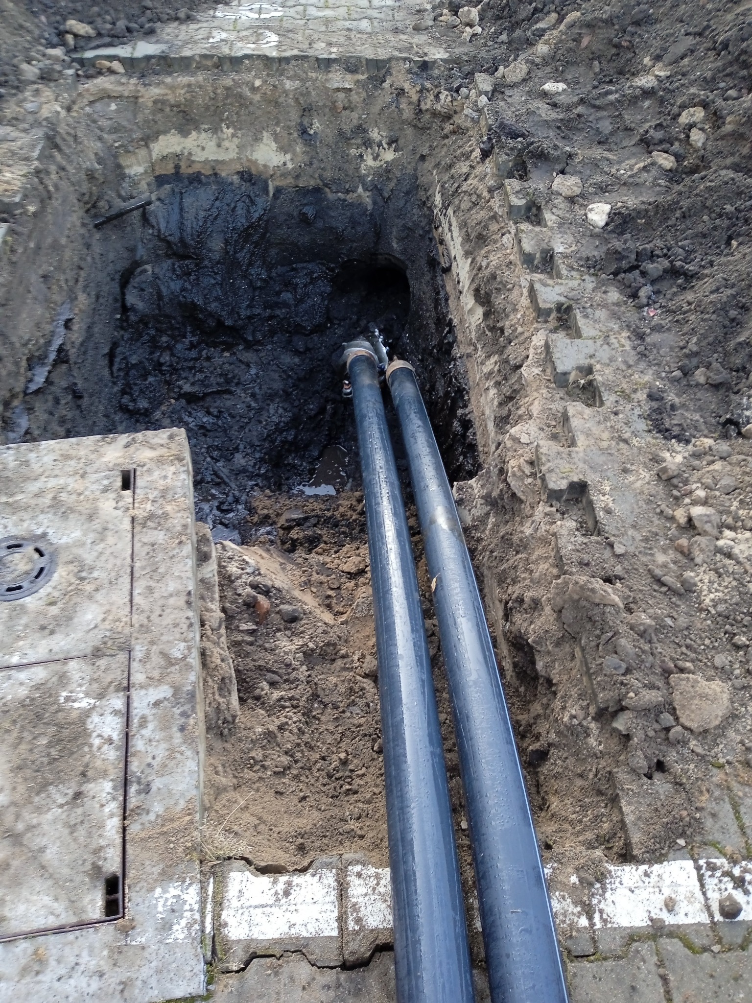 Dwie rury prowadzone w wykopie ziemnym, widoczny betonowy właz studzienki kanalizacyjnej po lewej stronie.
