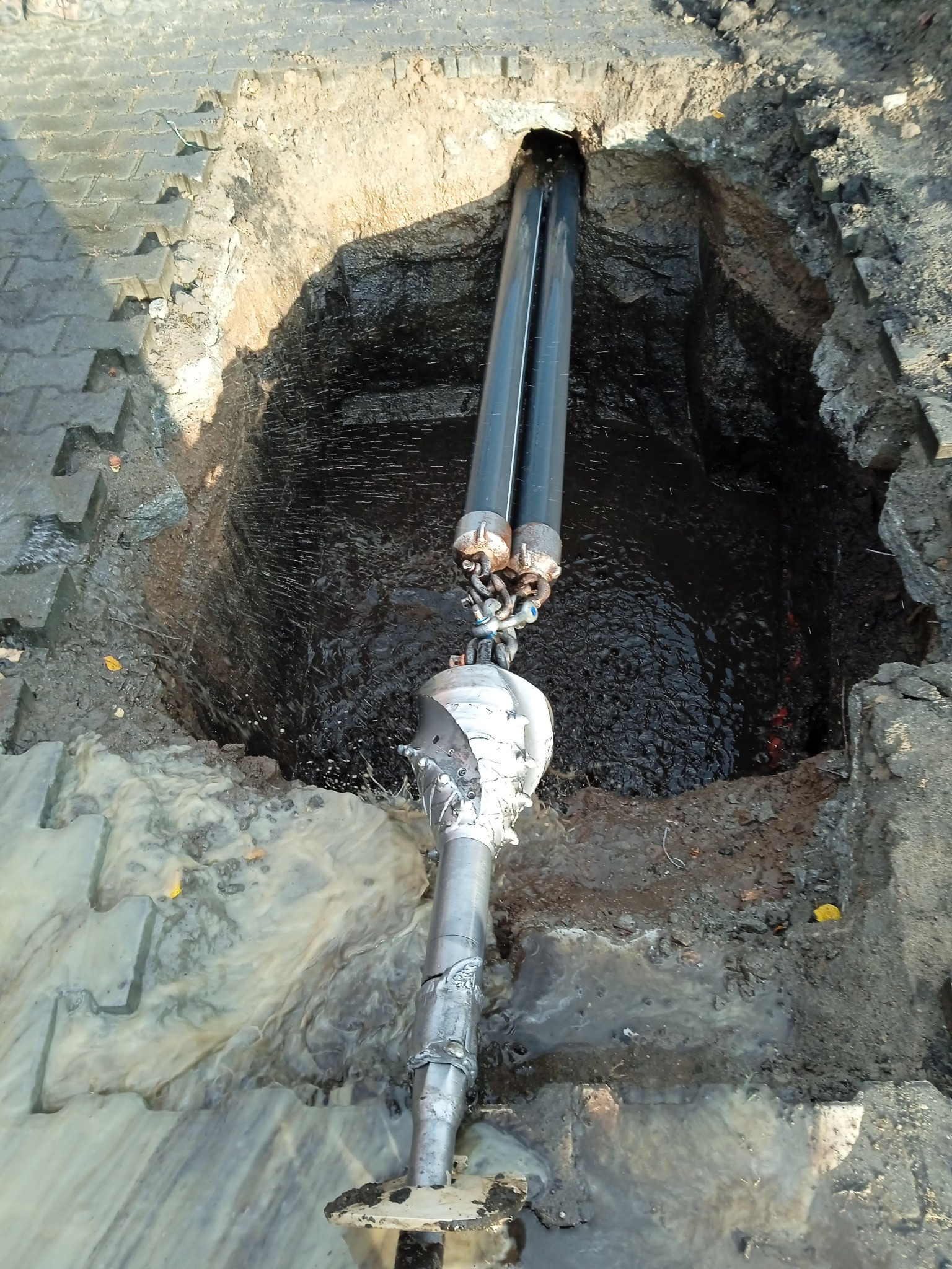 Wiertnica kierunkowa w wykopie, widoczne dwie rury i głowica wiertnicza w błocie, otoczenie z betonowej kostki brukowej i ziemi.