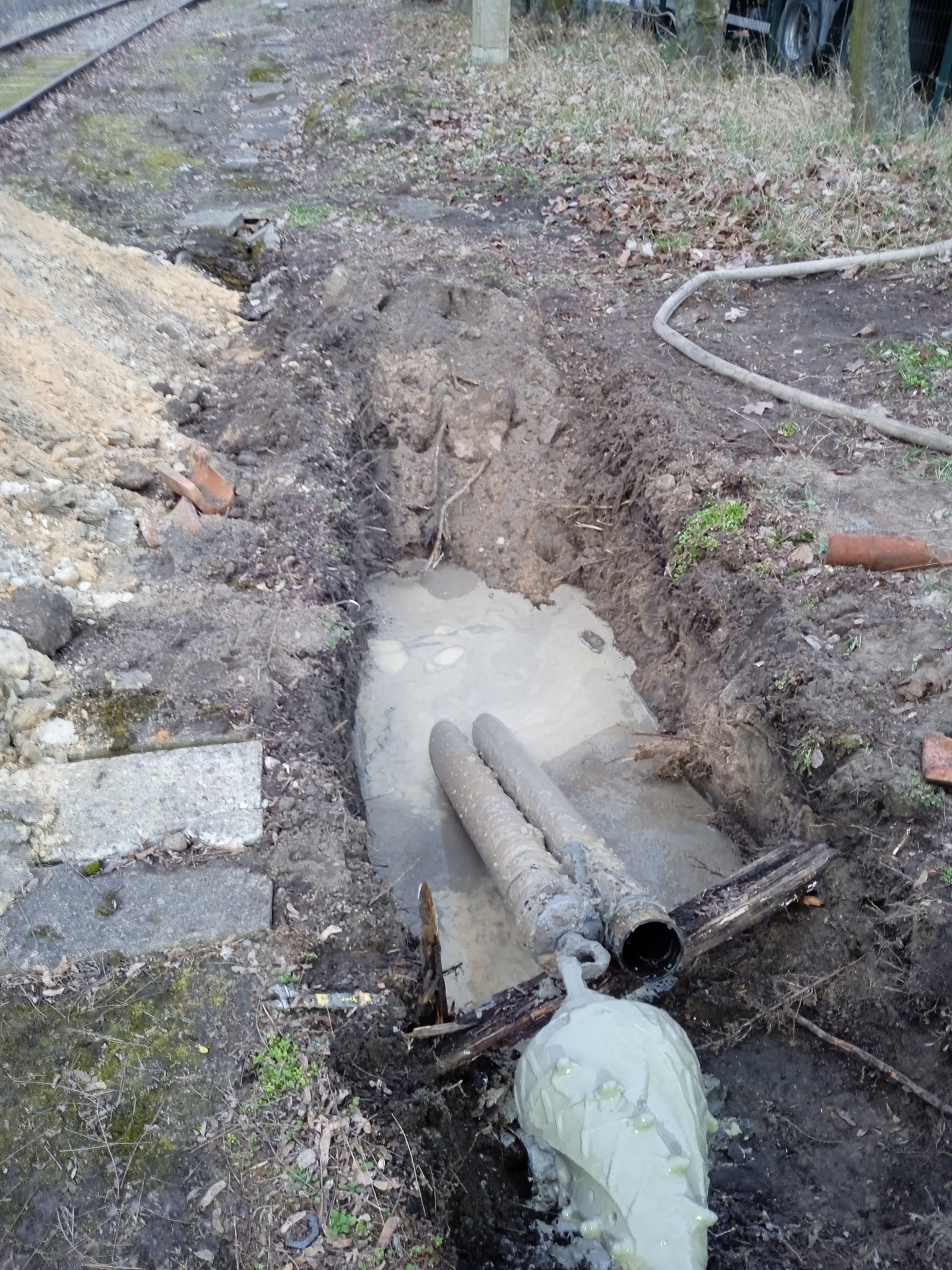 Dwa rurociągi w wykopie wypełnionym wodą, z widocznym elementem wiertniczym i błotnistym otoczeniem, w tle tor kolejowy.