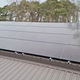 Panele fotowoltaiczne zamontowane na blaszanym dachu, widoczne uchwyty montażowe, w tle drzewa.