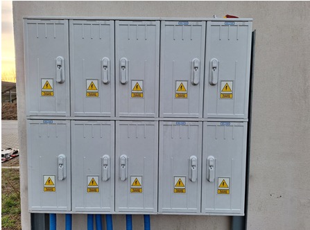 Zestaw dziesięciu szarych skrzynek elektrycznych zamontowanych na ścianie budynku w Galewicach, z widocznymi symbolami ostrzegawczymi i niebieskimi rurami osłonowymi u dołu.