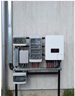 Zewnętrzna skrzynka elektryczna z licznikiem, rozdzielnicą i falownikiem fotowoltaicznym zamontowana na metalowej konstrukcji przy ścianie budynku. Widoczne kable i przewody. Szara ściana...