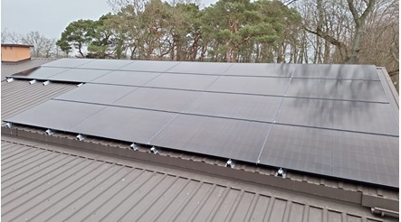 Panele fotowoltaiczne zamontowane na blaszanym dachu, widoczne uchwyty montażowe, w tle drzewa.