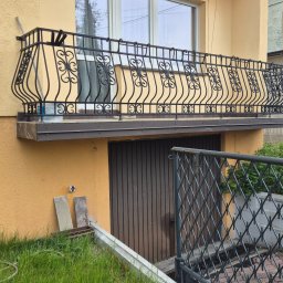 AGM AUTO-GARAŻ AGNIESZKA GAWROŃSKA - Żółta elewacja budynku z balkonem o ozdobnej, metalowej balustradzie. Widoczne okno i drzwi garażowe. Częściowo widoczny trawnik i ogrodzenie.