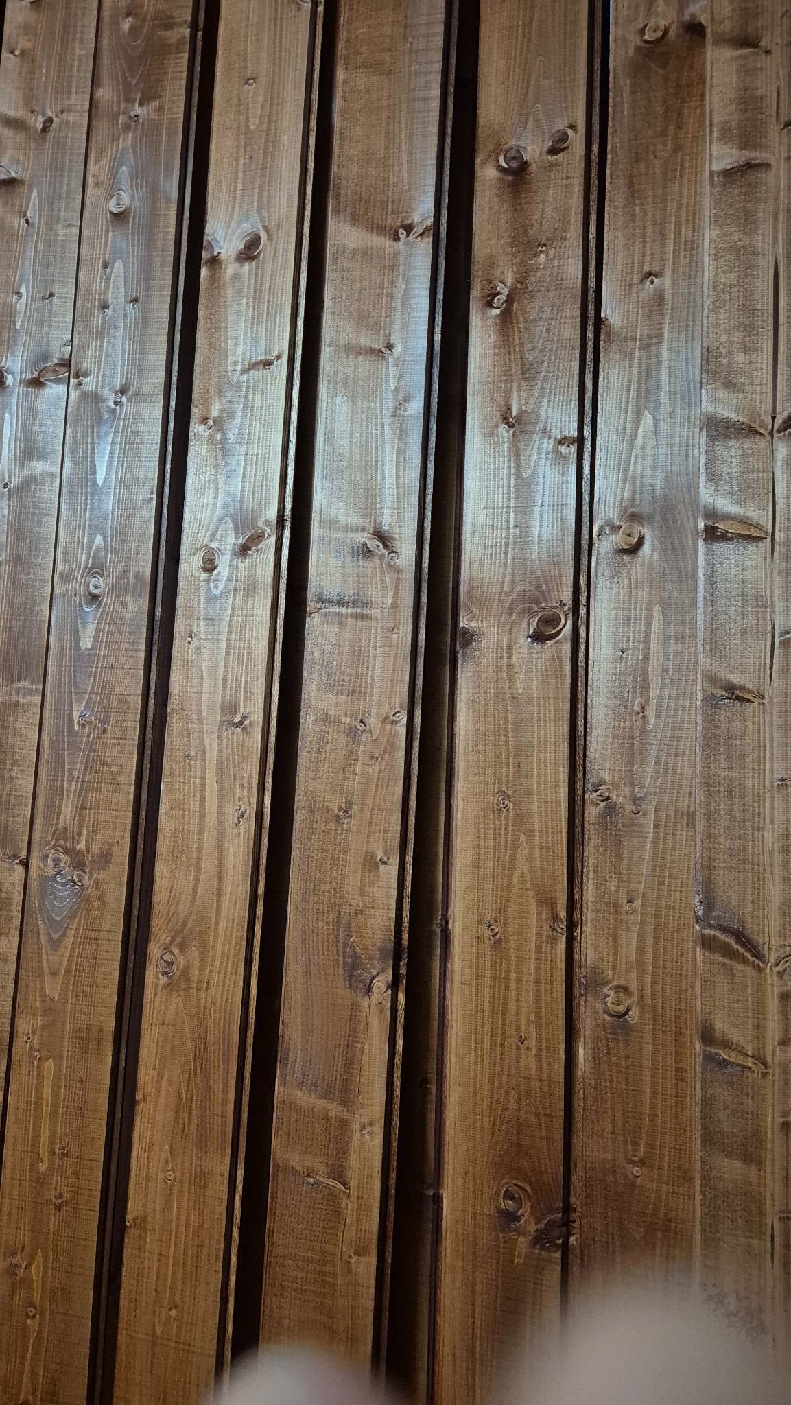 Lakierowane deski boazeryjne w odcieniach brązu, pionowo ułożone, z widoczną strukturą drewna i ciemniejszymi, wąskimi listwami pomiędzy deskami.