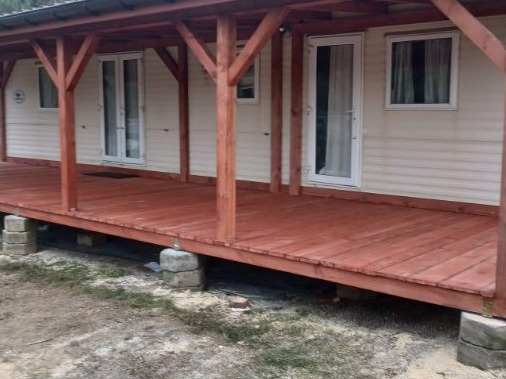 Zadaszony taras drewniany z bejcowanymi deskami w odcieniach czerwieni, wsparty na betonowych blokach. Widoczne białe drzwi i okno w tle.