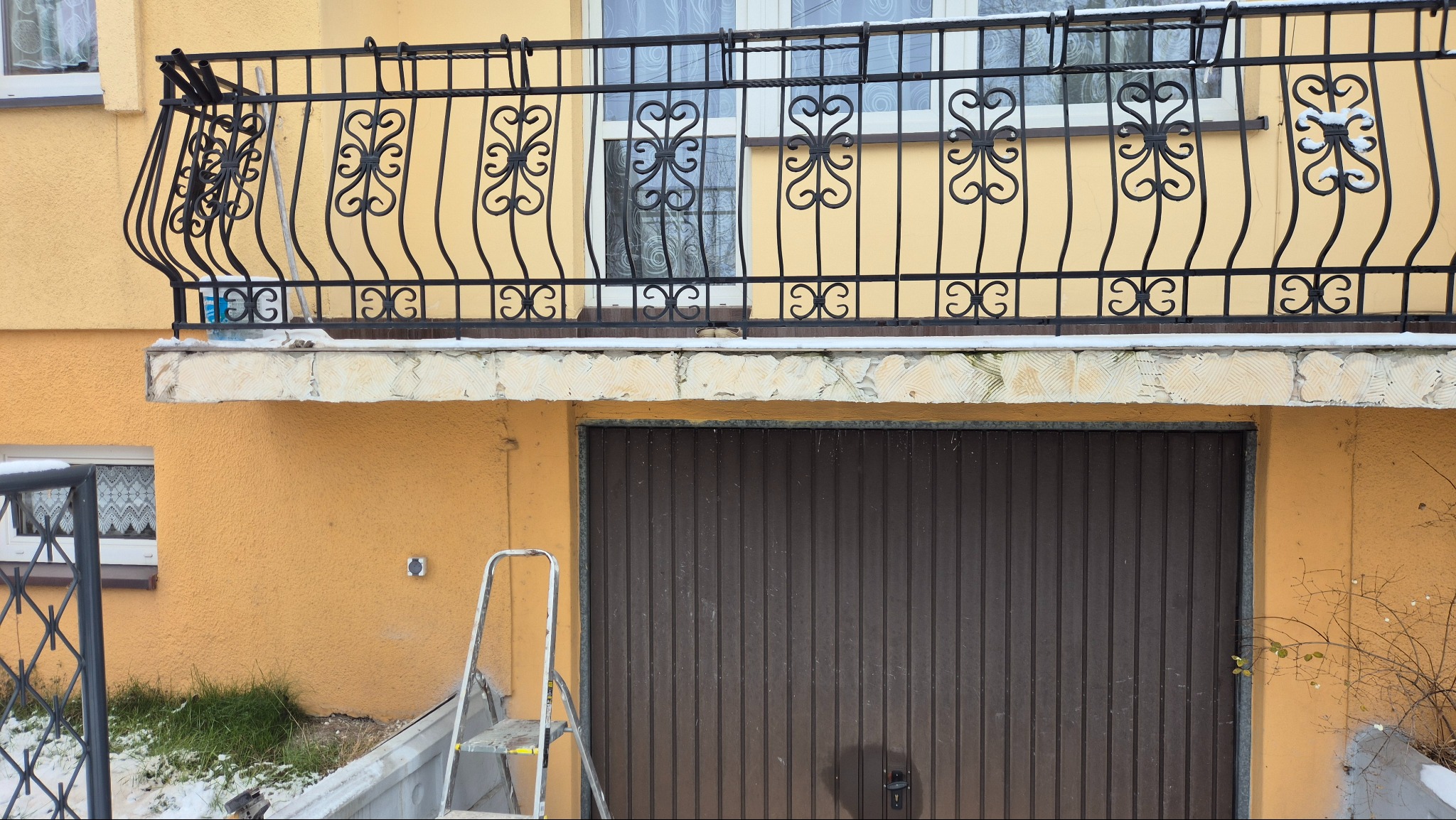 Żółty budynek z balkonem z ozdobną, czarną balustradą. Widoczna drabina oparta o ścianę i brama garażowa. Fragmenty śniegu na balkonie i trawie.