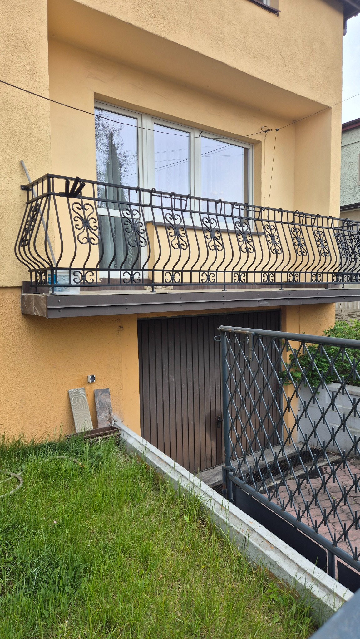 Żółta elewacja budynku z balkonem o ozdobnej, metalowej balustradzie. Widoczne okno i drzwi garażowe. Częściowo widoczny trawnik i ogrodzenie.