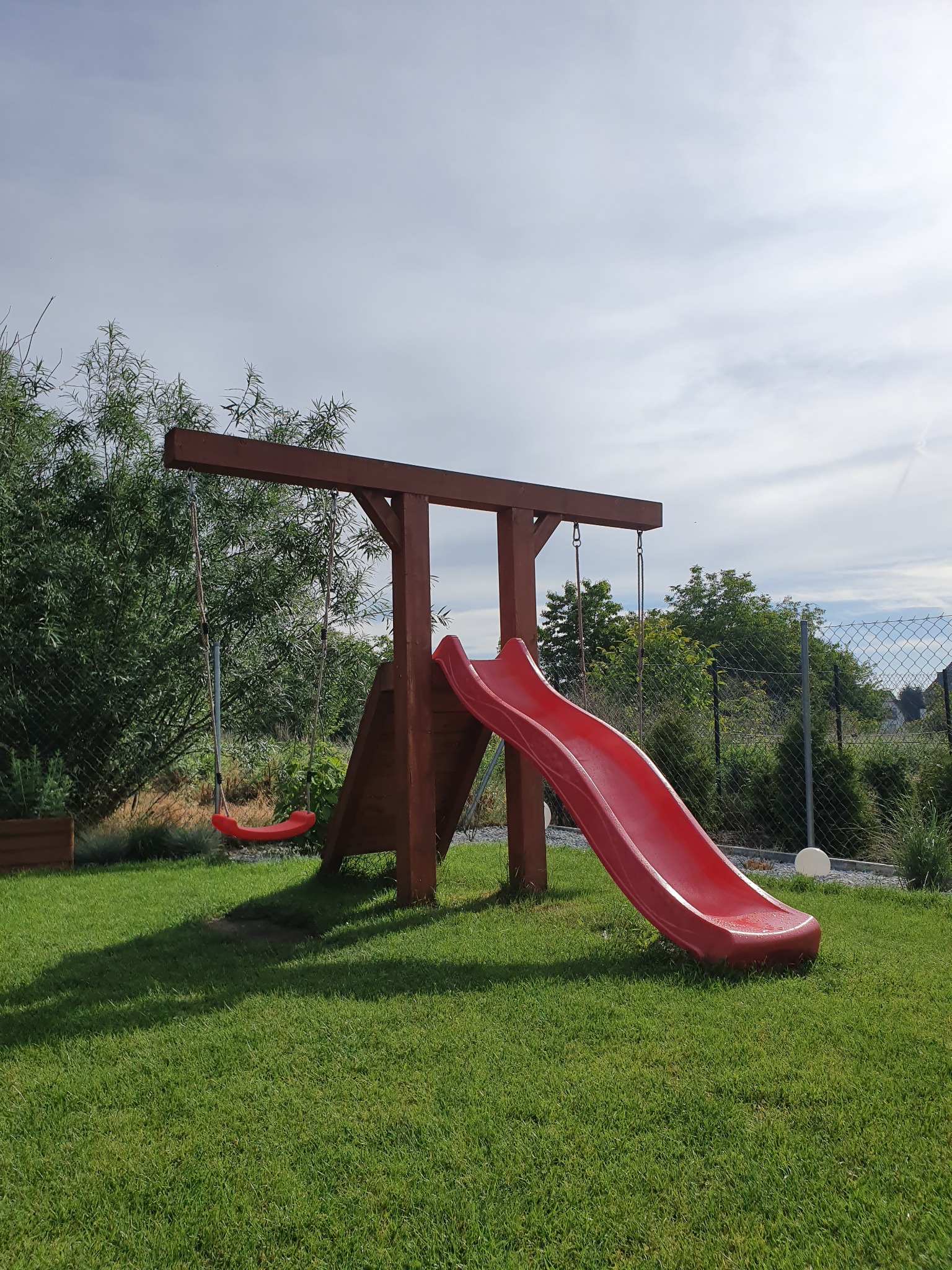 Plac zabaw z drewnianą konstrukcją, czerwona zjeżdżalnia i huśtawka na tle zielonego trawnika i drzew