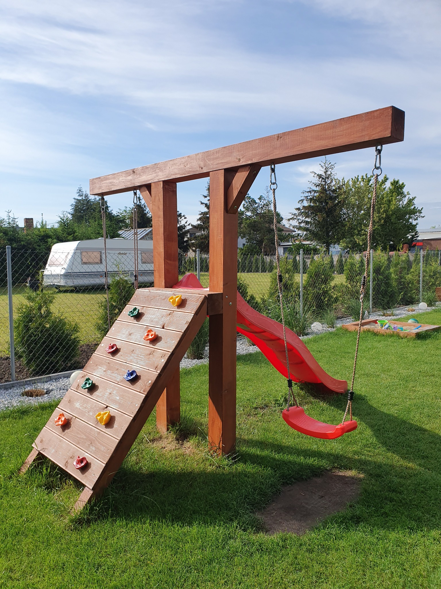 Drewniany plac zabaw z czerwoną zjeżdżalnią i huśtawką na tle zadbanego trawnika i przyczepy kempingowej.